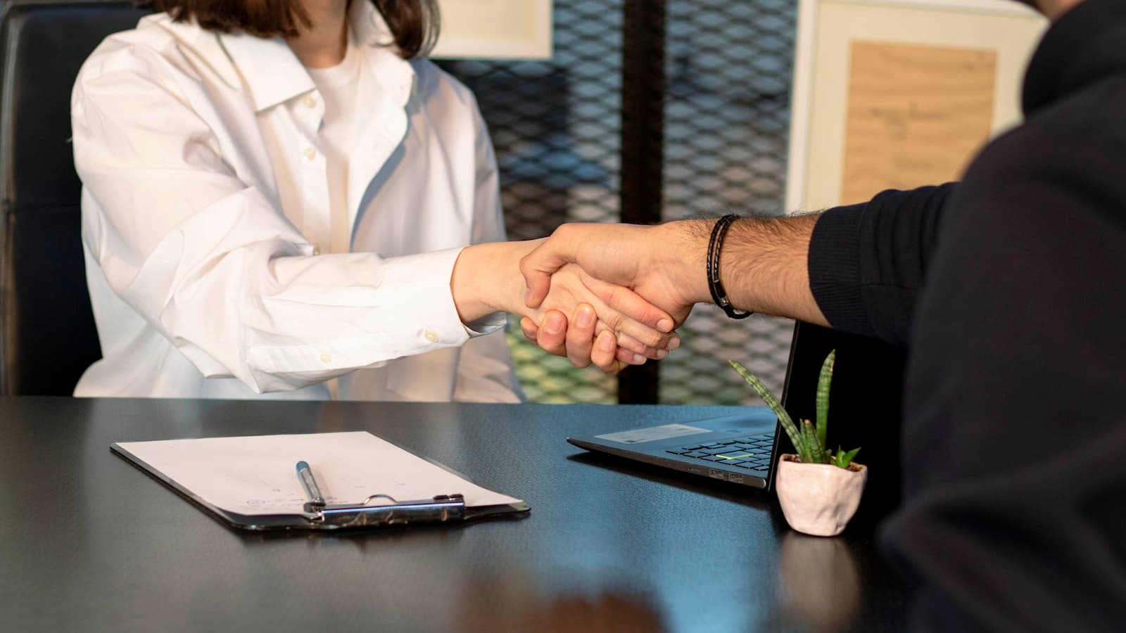 A handshake over a desk with a laptop and clipboard of documents.