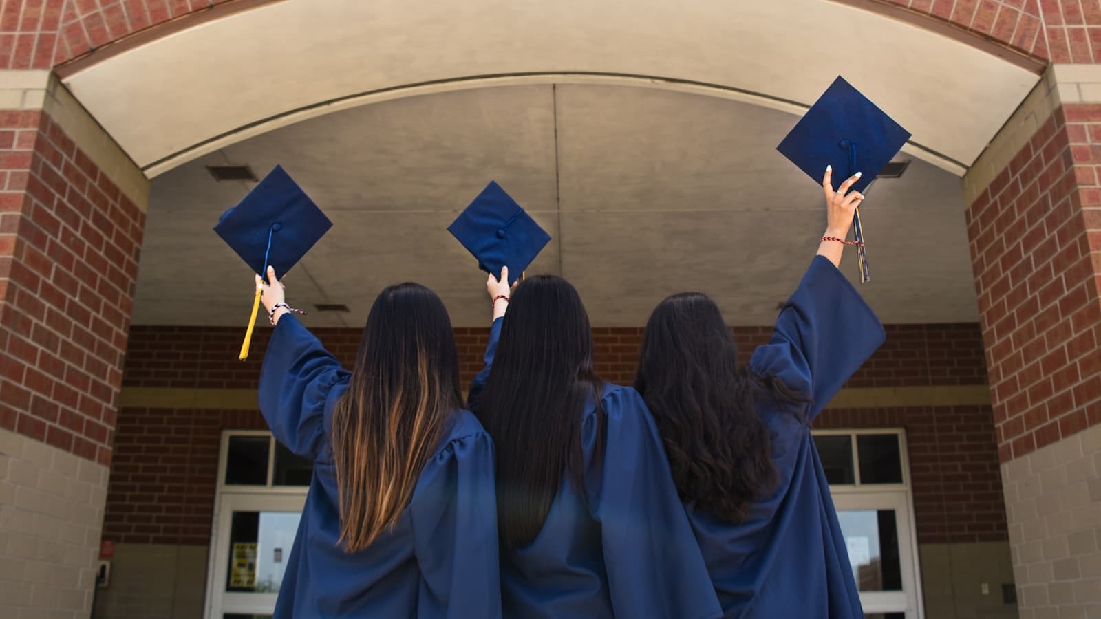 Three graduates in navy caps and gowns stand with their backs to the camera, raising their mortarboards overhead in front of a brick building archway.