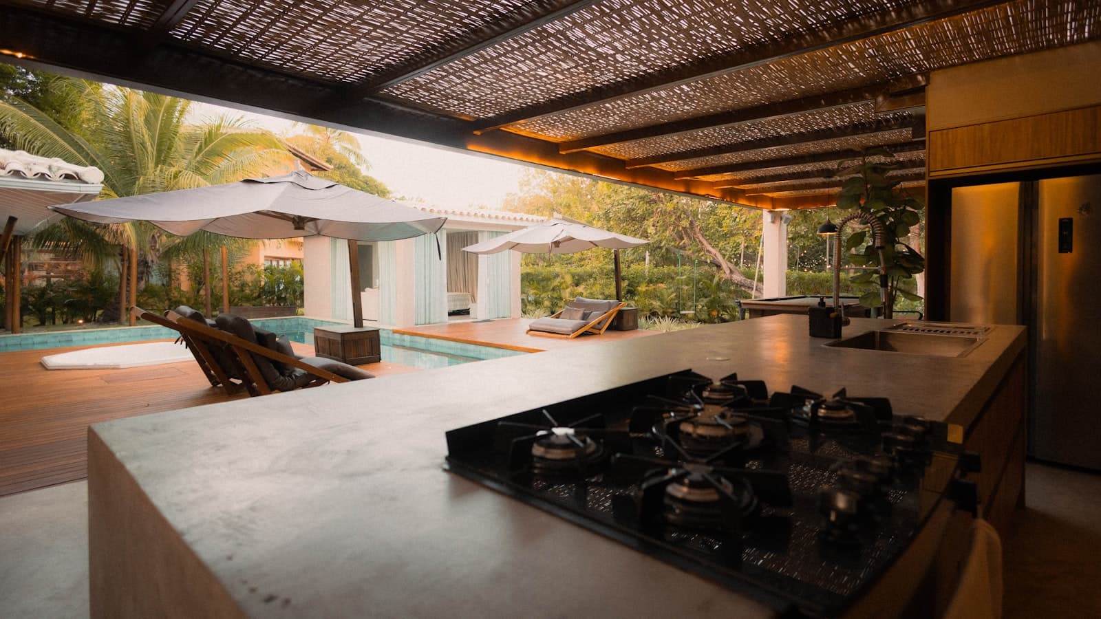 A concrete outdoor kitchen island with a built-in gas stovetop and sink sits under a woven canopy roof, overlooking a pool deck with umbrellas and lounge chairs at dusk.