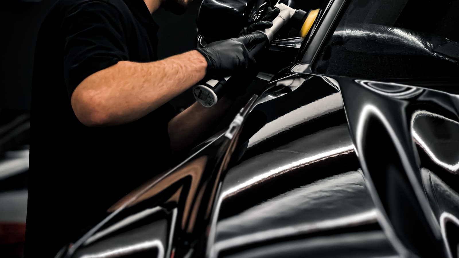 A man in a dark garage waxing and polishing the hood of a black car by hand.