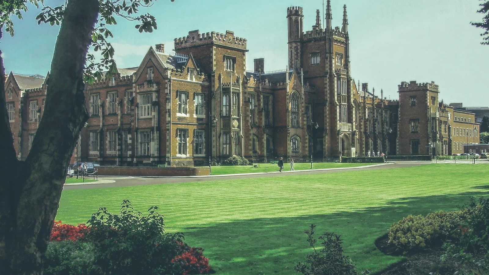 A Gothic-revival collegiate stone building with towers behind a lawn, two unidentifiable distant pedestrians, no legible text or crest visible.
