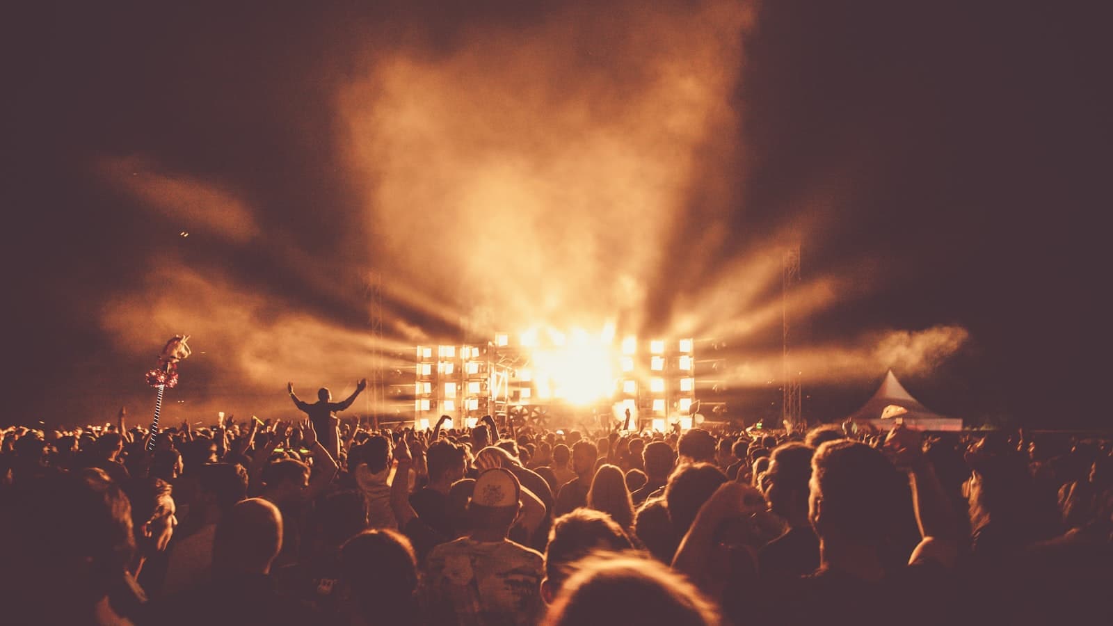 A concert crowd silhouetted with raised arms facing a brightly lit stage; no identifiable faces or readable text.