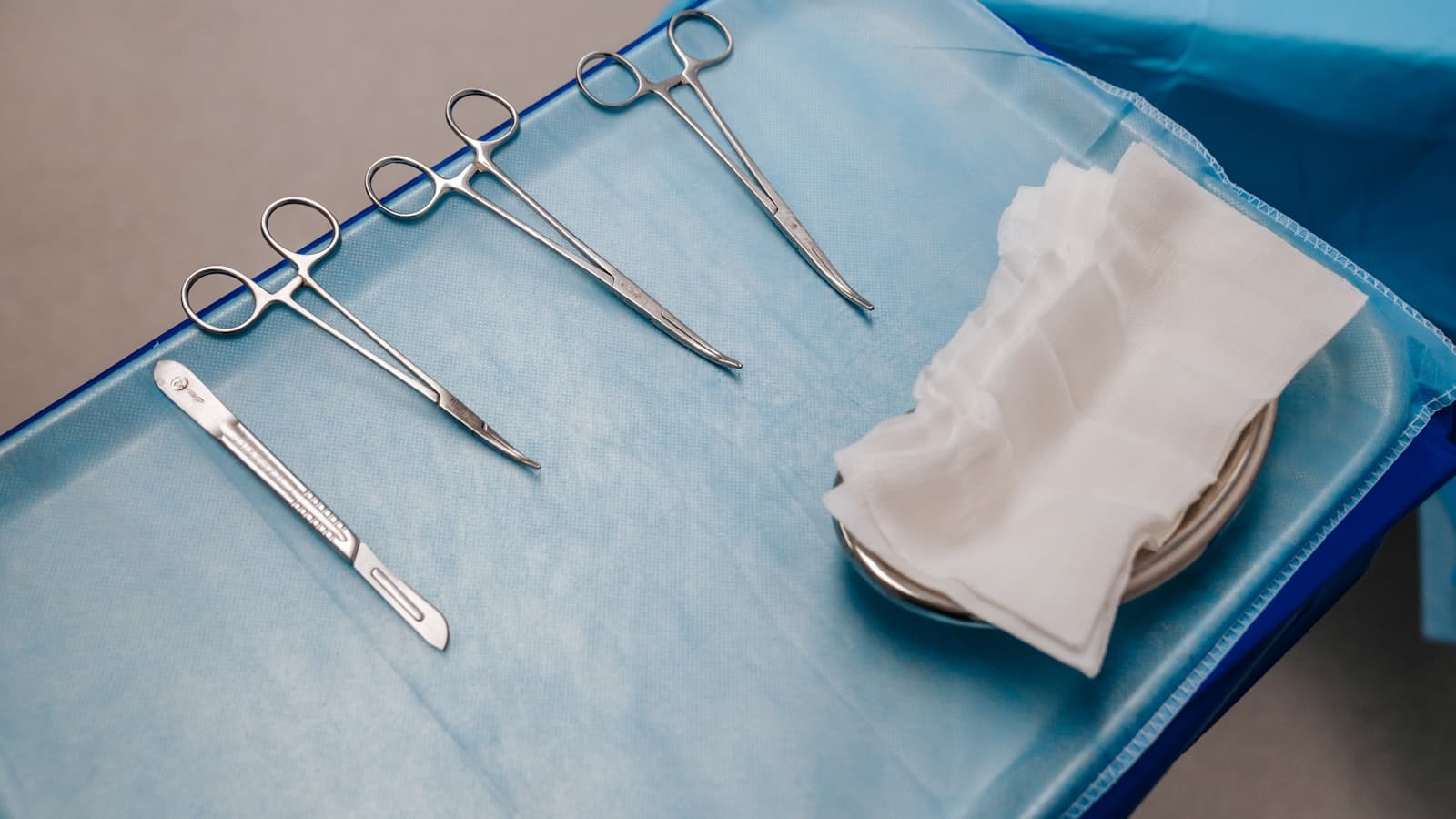 A sterile tray of surgical instruments (scalpel, clamps, gauze) on blue medical drape, no people or readable text.