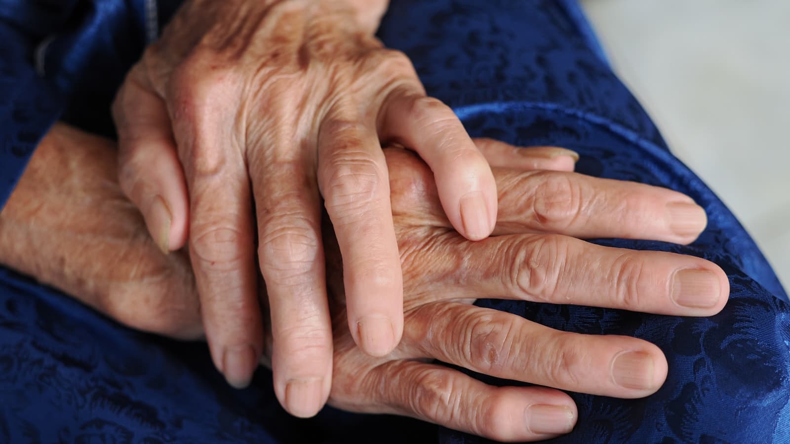 Close-up of an elderly person's wrinkled hands clasped together resting on blue fabric, no face, text, or logos visible.