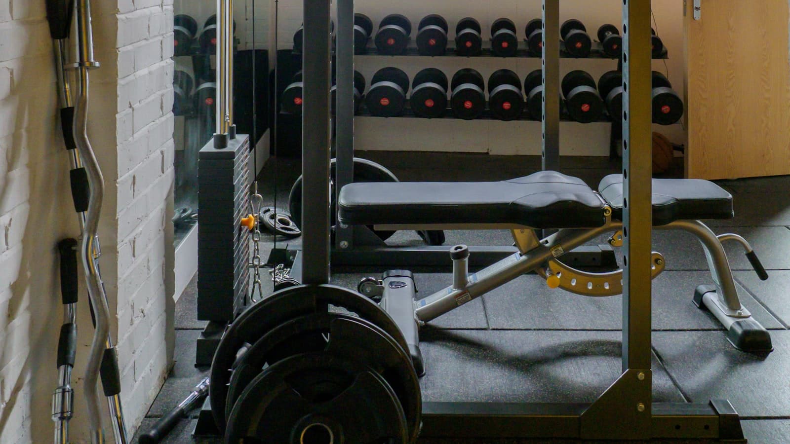 A converted garage home gym with an exposed brick wall, a rack of dumbbells, a flat weight bench, and a loaded barbell on a squat rack.