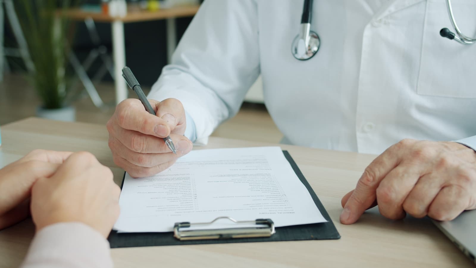 Close-up of a doctor's hand writing on a clipboard with the patient's hand in frame.