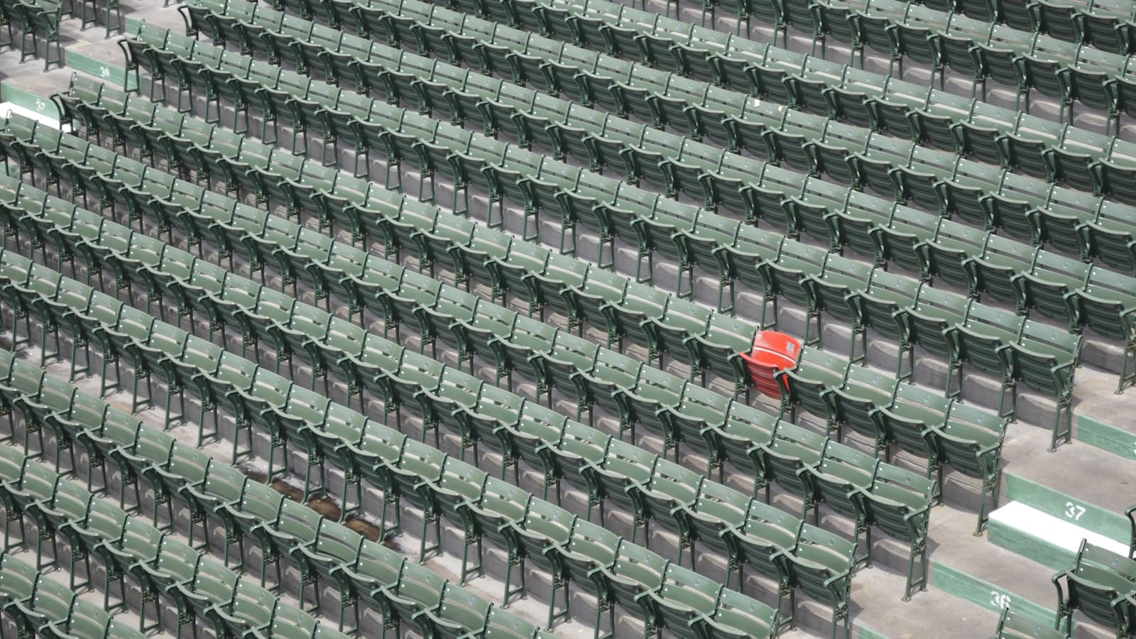 Rows of empty green stadium seats with one lone red seat marked among them; only small row numbers visible, no logos.