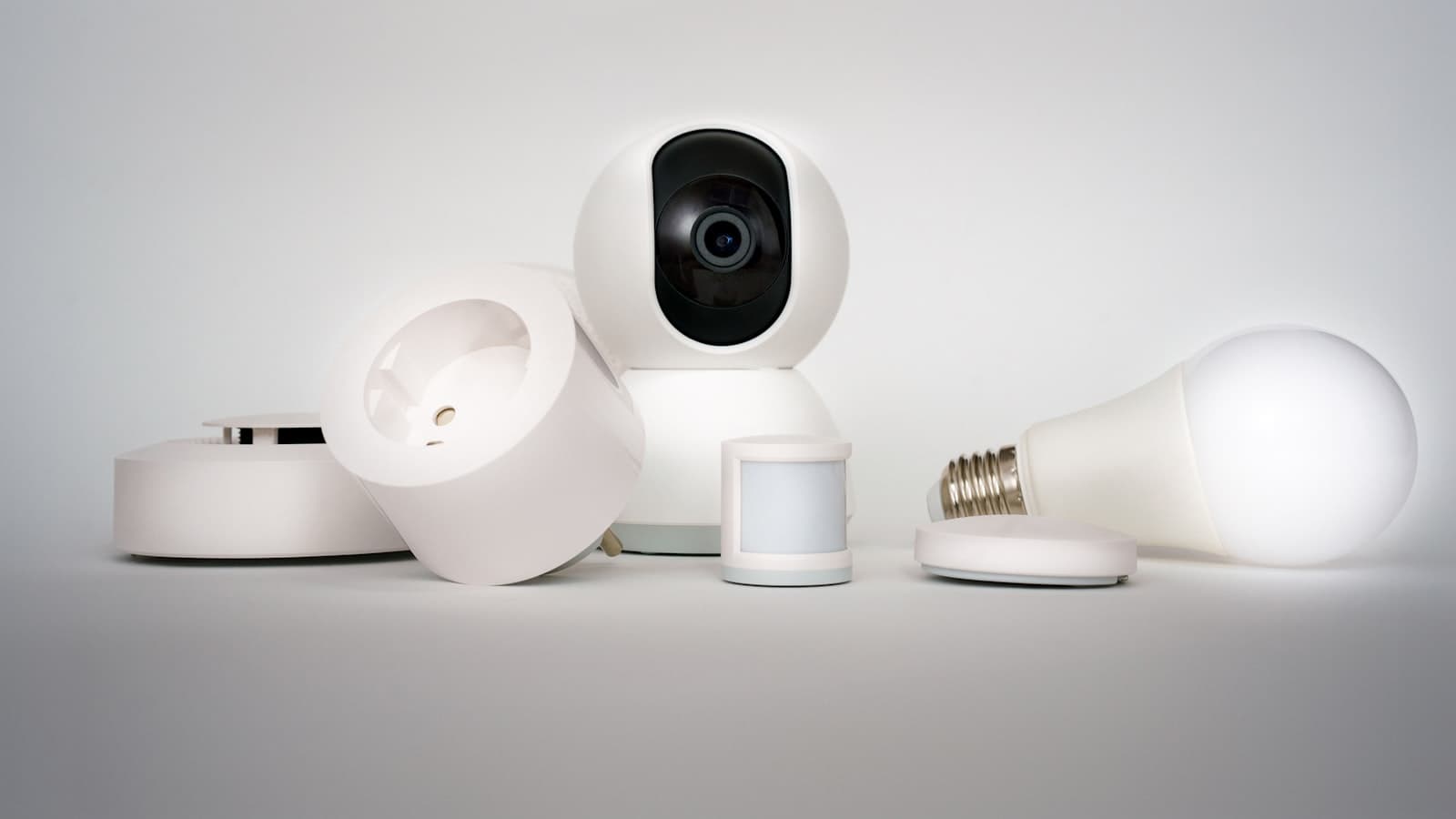 A clean studio arrangement of white smart home devices - a camera, motion sensor, smart plug, smoke detector, and smart bulb; no faces or logos.