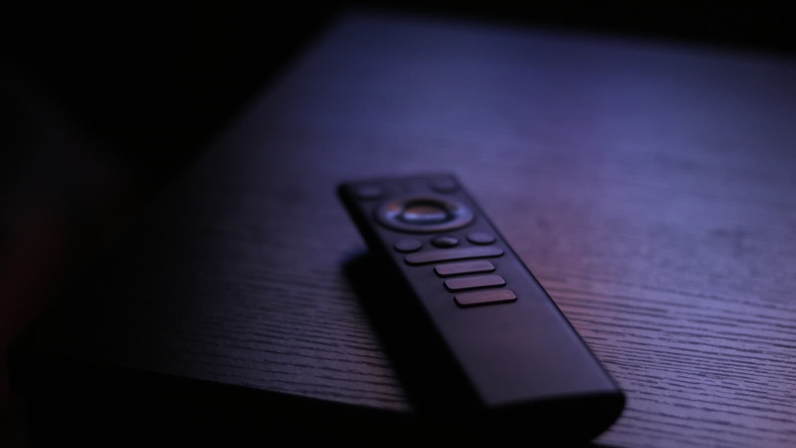 A TV remote control resting on a dark wooden table in dim, moody lighting, with buttons too dark to read and no logos or people.