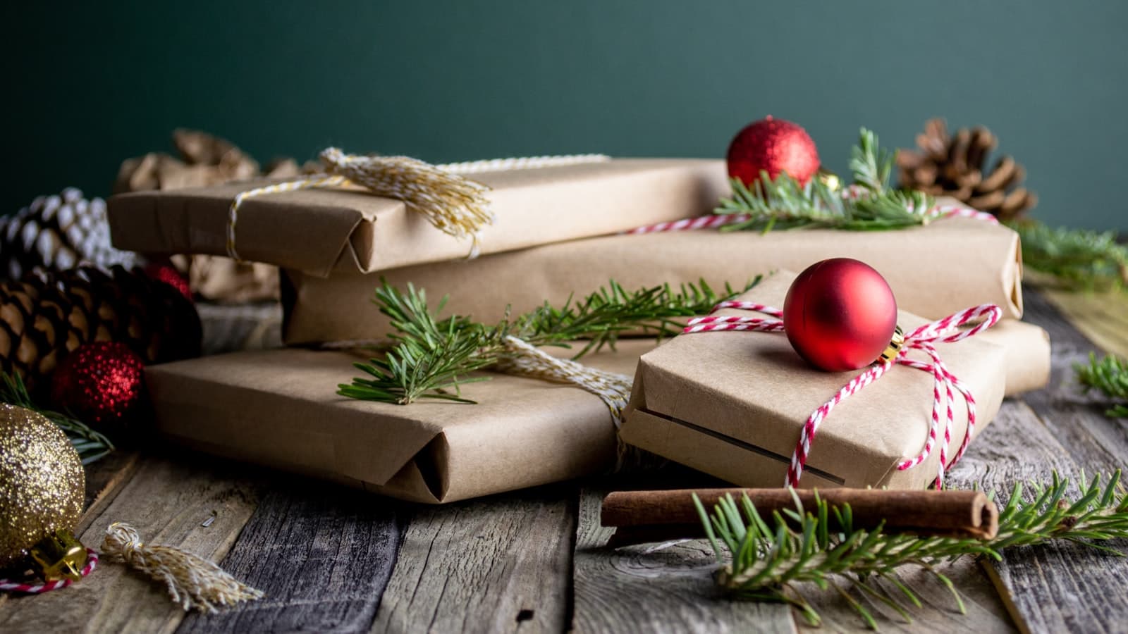 Several kraft-paper-wrapped gift boxes tied with string and decorated with red ornaments and pine sprigs, arranged on a table against a dark green background - no people or text.