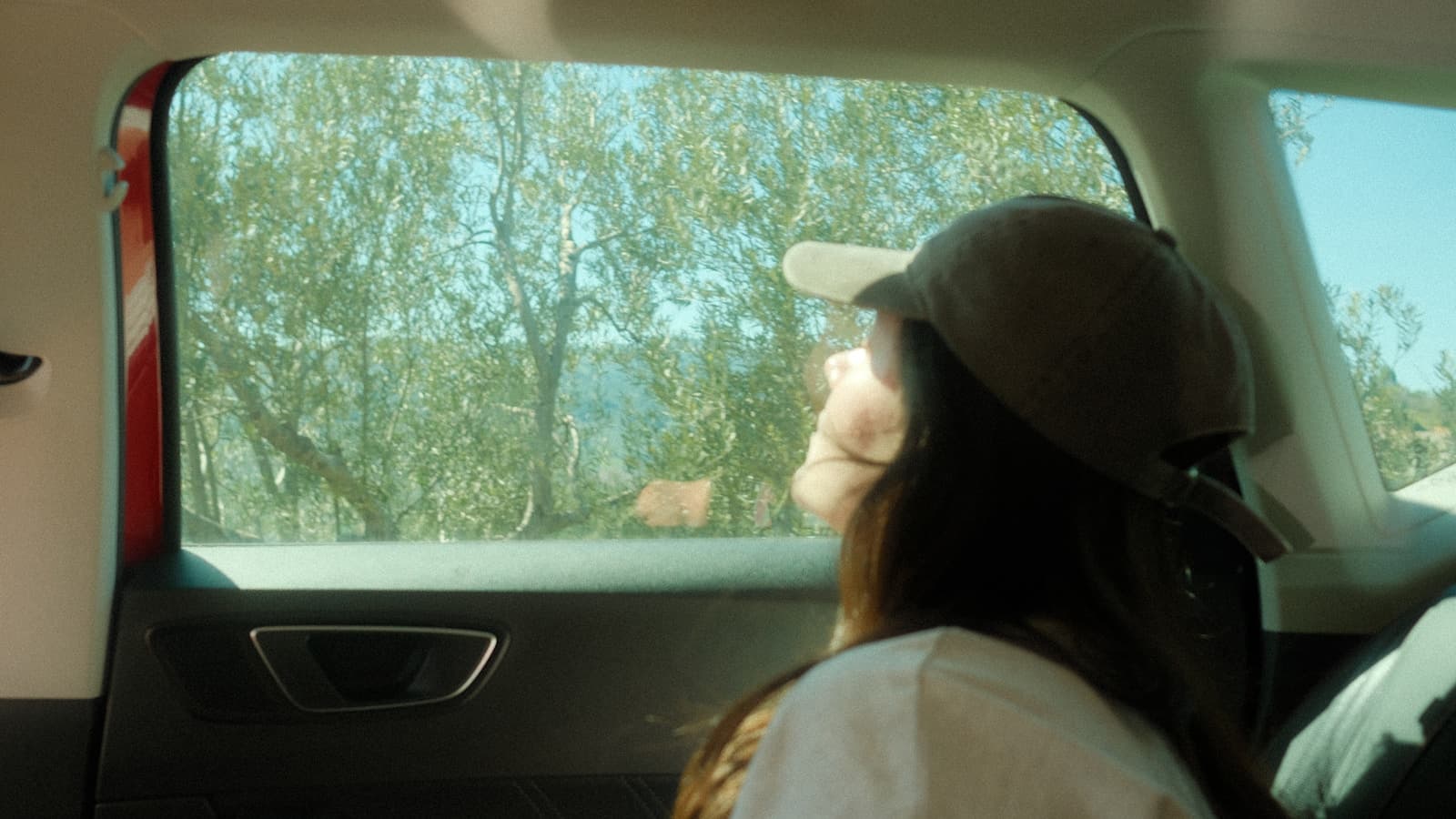 A woman in a cap looking out the back-seat car window at passing trees.