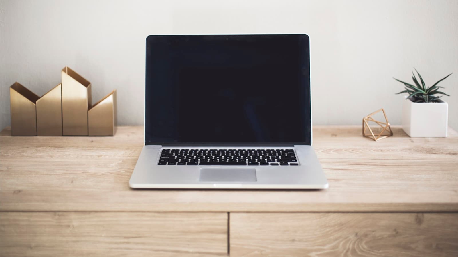A laptop with a closed dark screen on a wooden desk with a small plant; no logos or text visible.