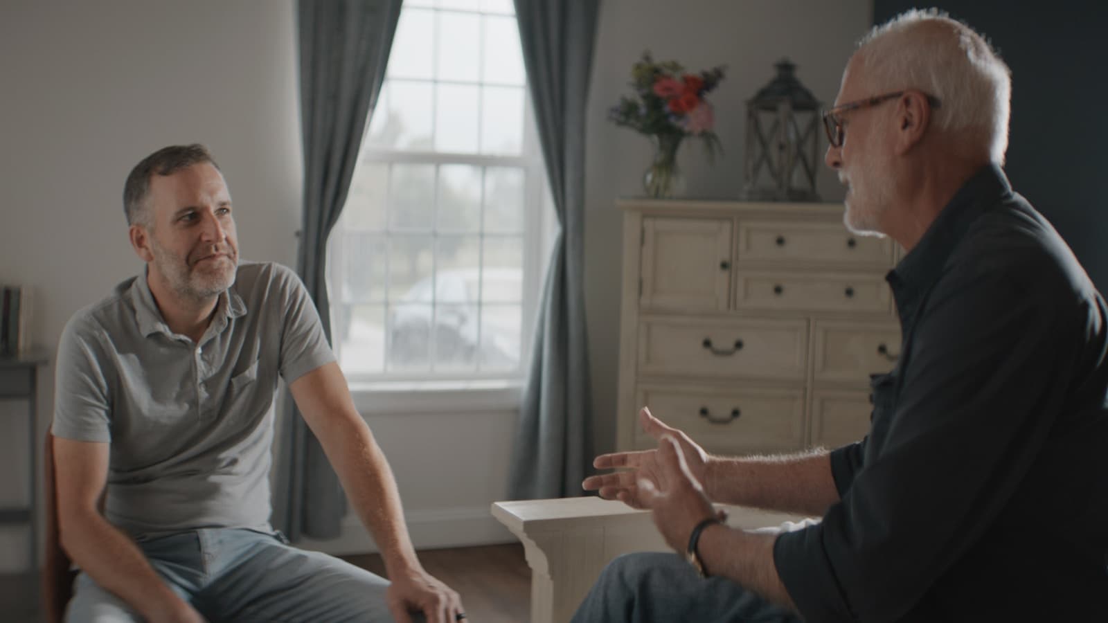 An older man and younger man in a warm, serious bedroom conversation, mentorship tone.