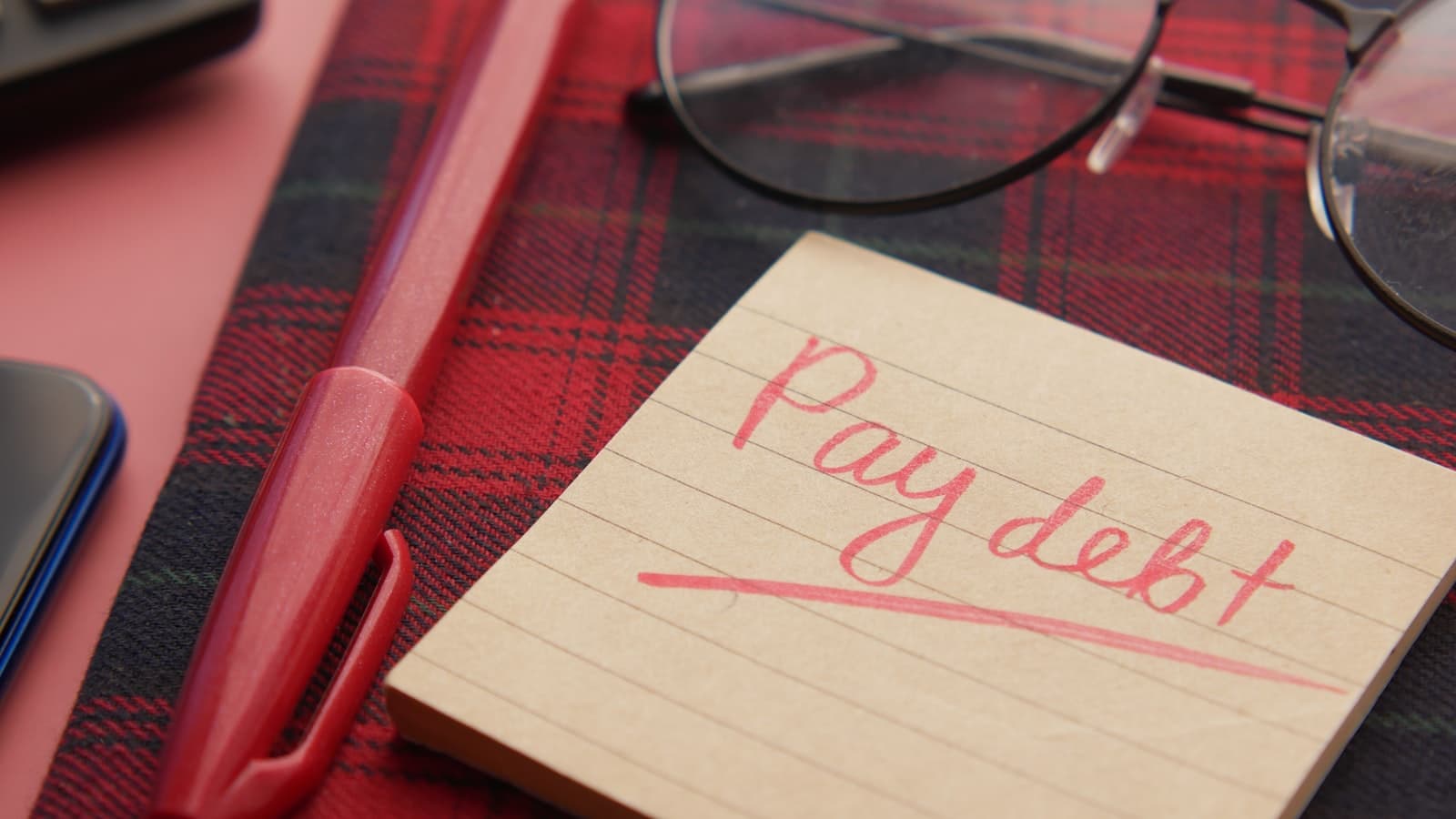 A handwritten note reading 'Pay debt', underlined, next to a pen and glasses