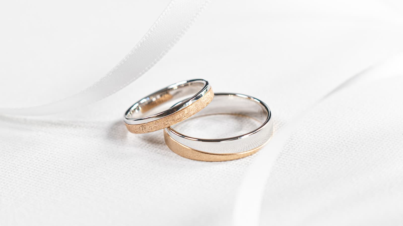 Two wedding rings, one silver and one gold, resting together on white fabric with a ribbon