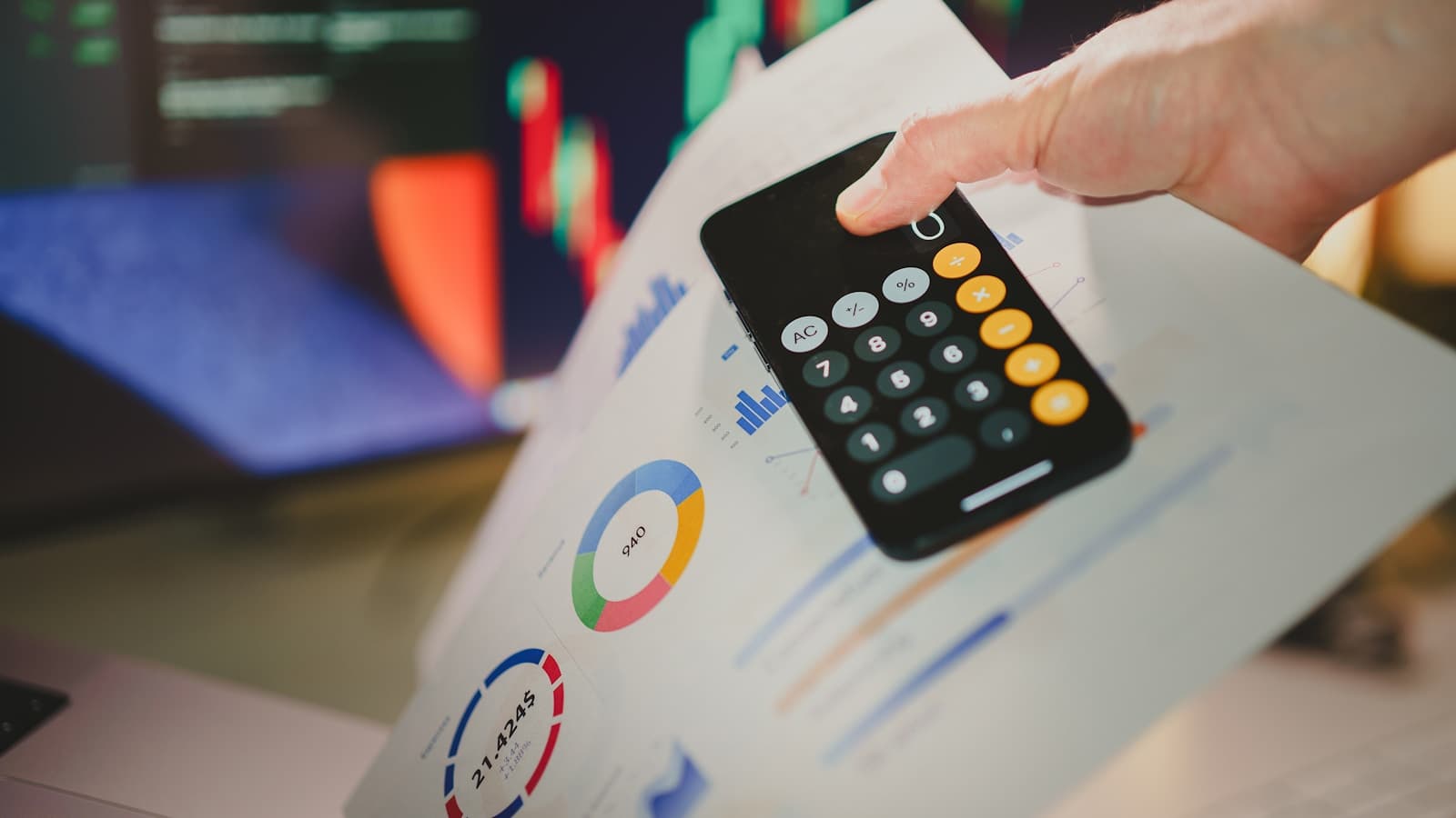 A hand holding a calculator app on a phone over blurred generic charts.