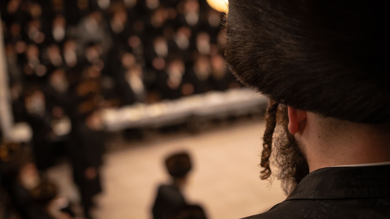 A man in formal dress seen from behind, looking out over a large seated family and community gathering.