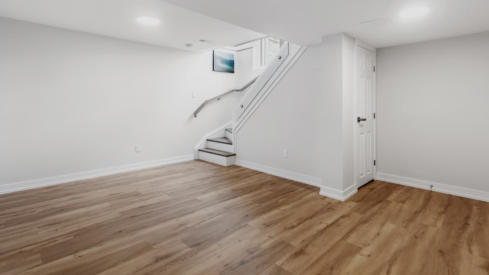 A freshly finished basement room with light wood flooring, white walls, and a staircase, no people or readable text.