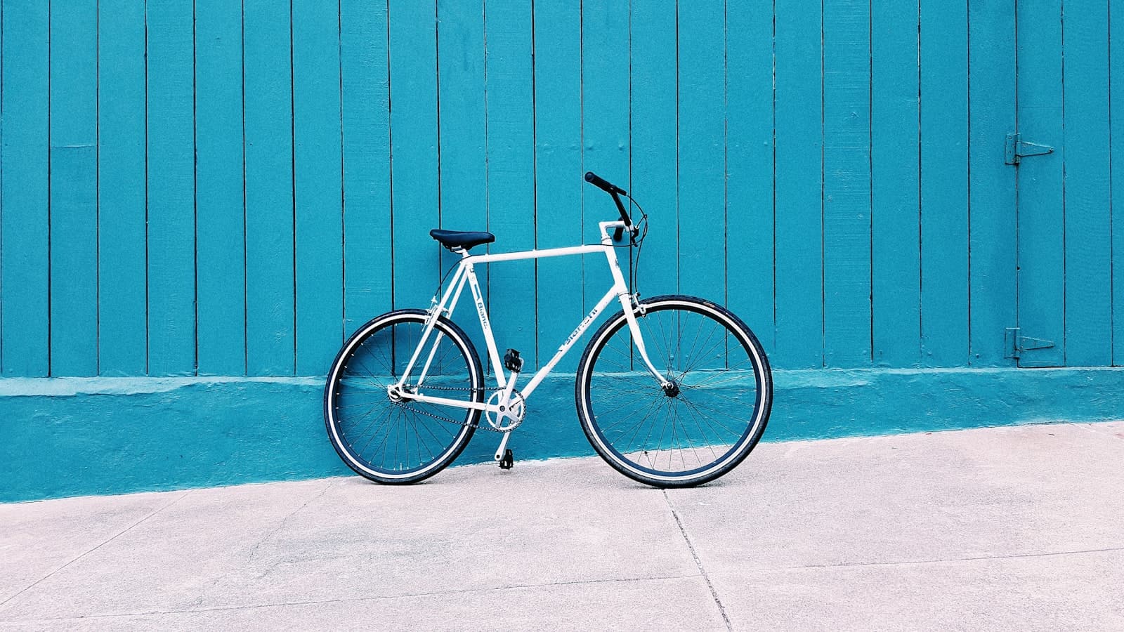 A white bicycle leaning against a teal wooden wall on a sidewalk, with no people and no legible branding.