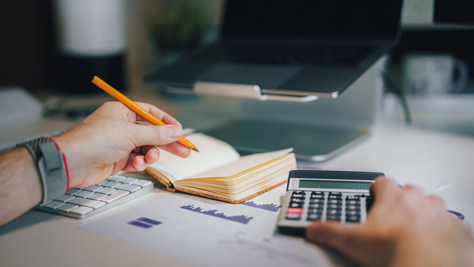 Close-up of hands with a pencil, notebook, and calculator over a printed financial bar chart.