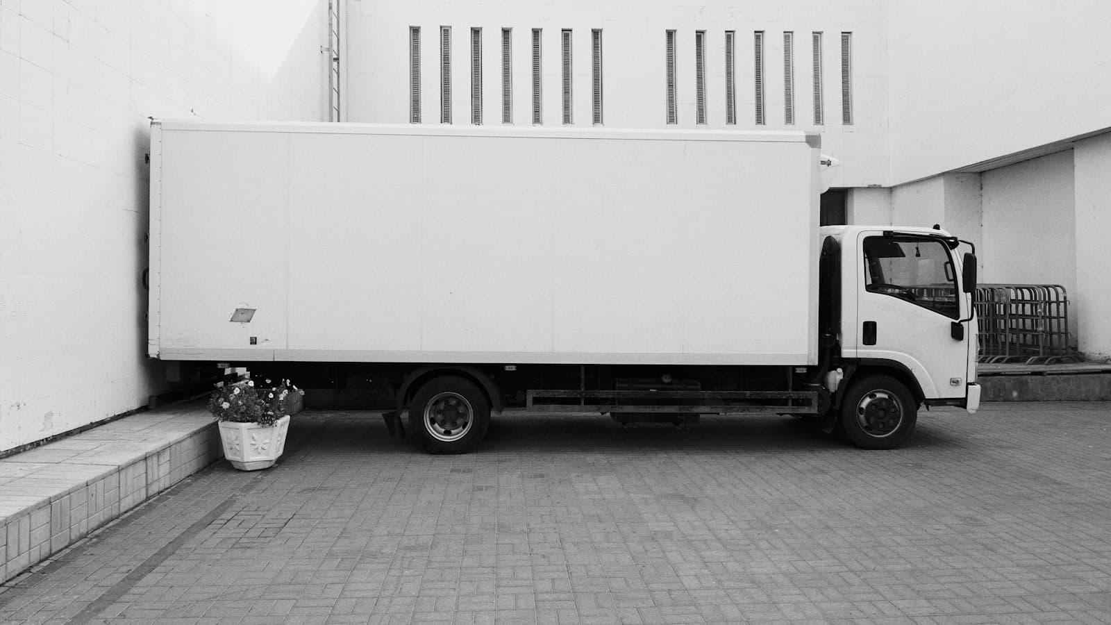 A black-and-white side view of a plain white unmarked box truck parked in an alley, with no people, text, or plate visible.