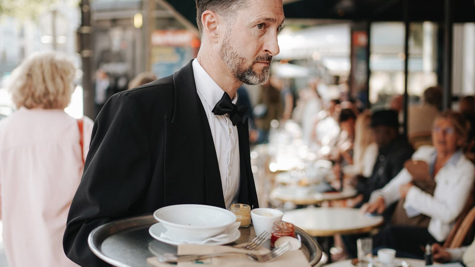 A man in a tuxedo carrying a serving tray outdoors at an upscale venue.