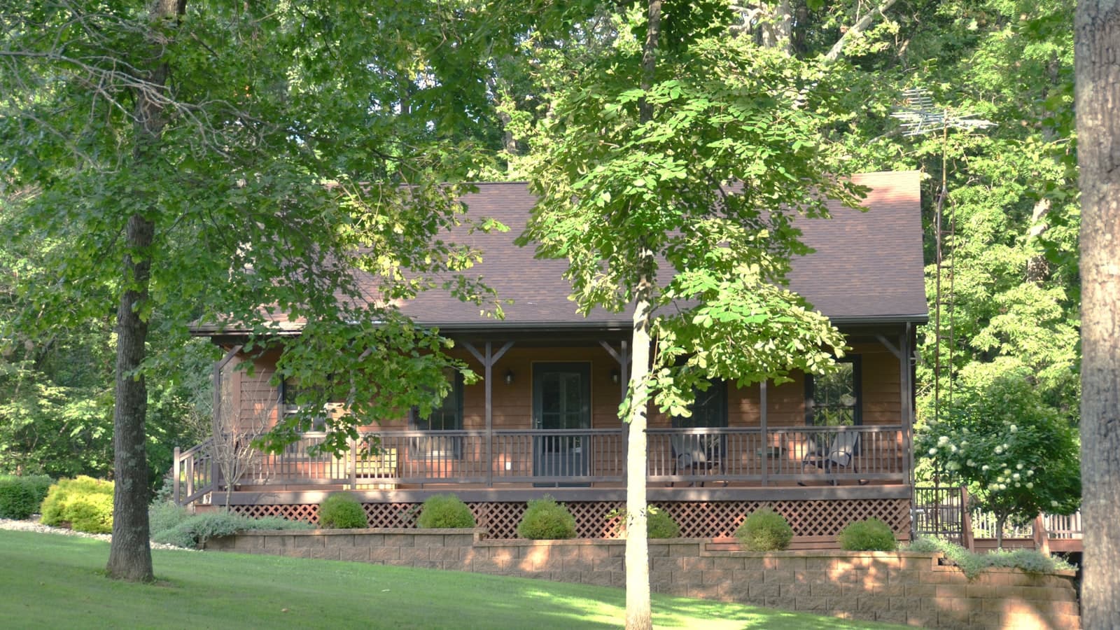 A wooden cabin with a covered porch and lattice skirting, surrounded by dense green forest.