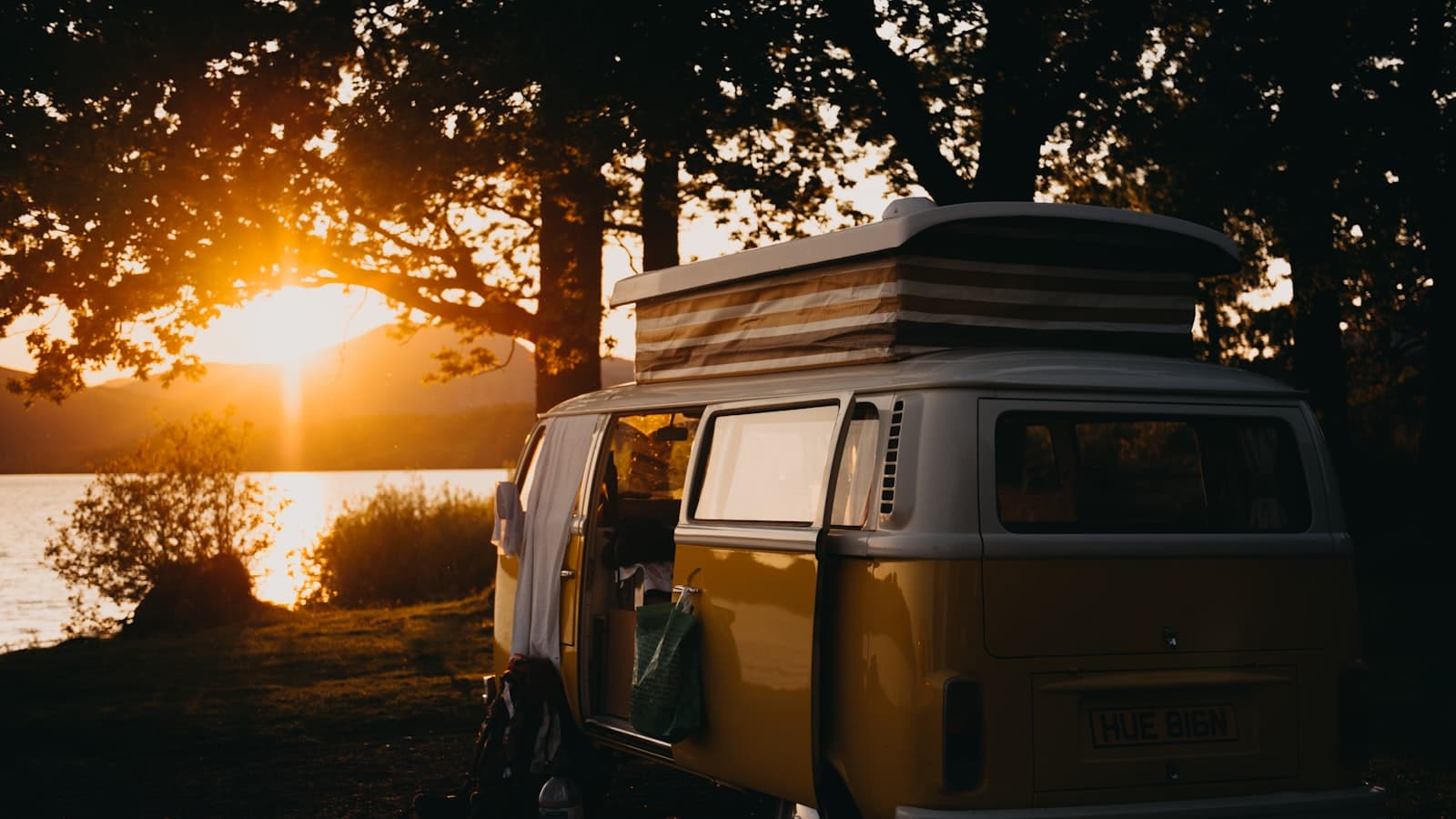 A green VW-style camper van with its side door open revealing a wood-paneled interior, parked by a lake at sunset with no visible plate or people.