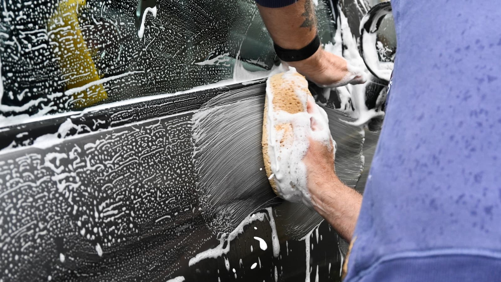 A hand with a sponge working thick foam across a dark car door panel.