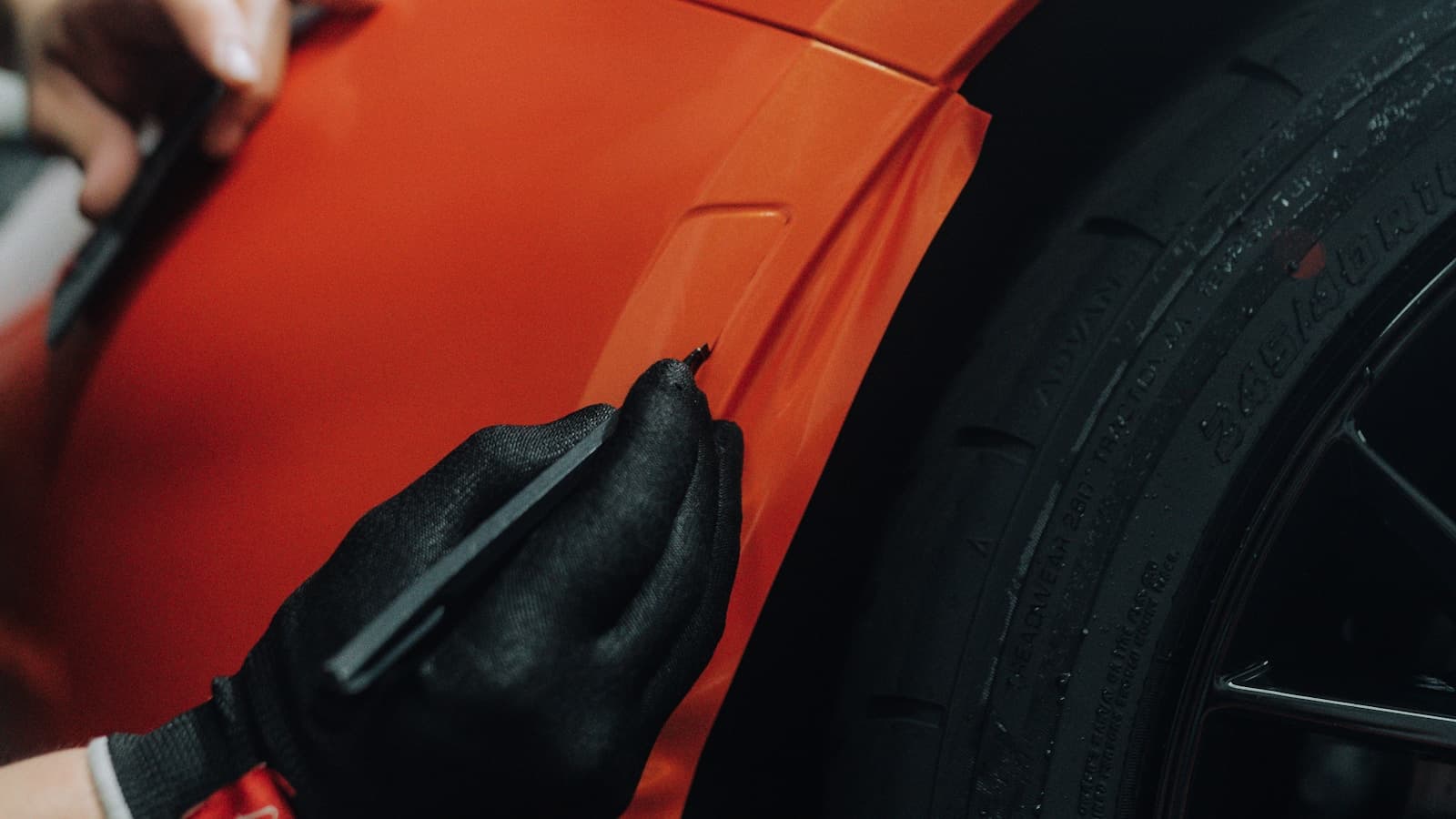 Close-up of a gloved installer squeegeeing an orange vinyl wrap film onto a car's rear fender.
