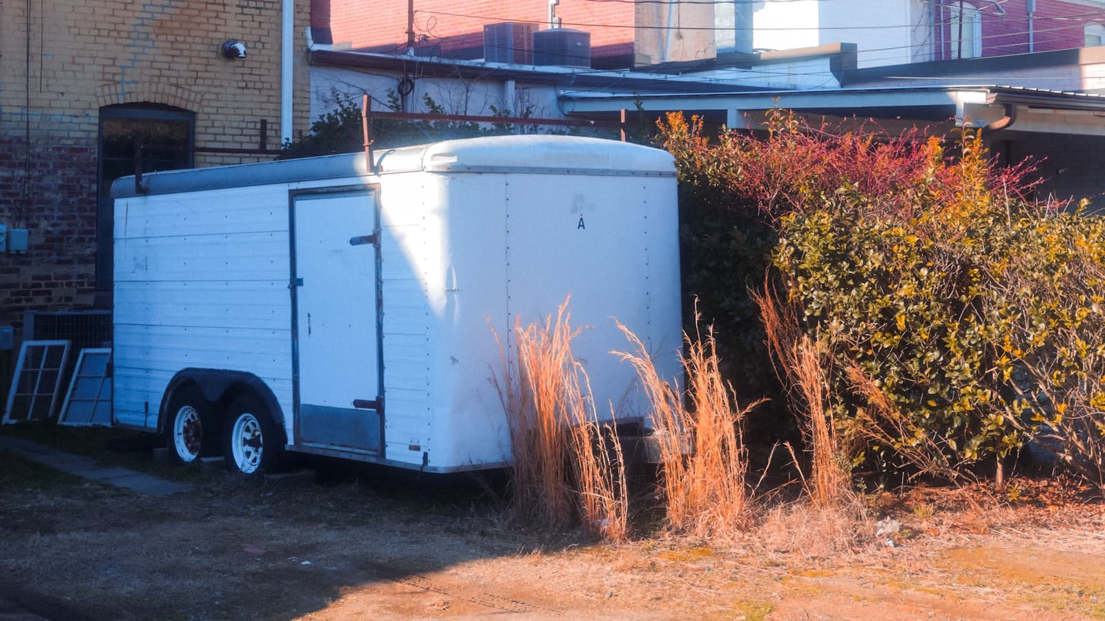 A plain white enclosed cargo trailer parked in a backyard alley beside a brick building, with no people, logos, or readable plate.