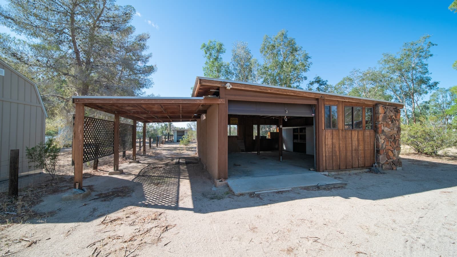 A wood-framed open carport structure attached to a house on a rural property.