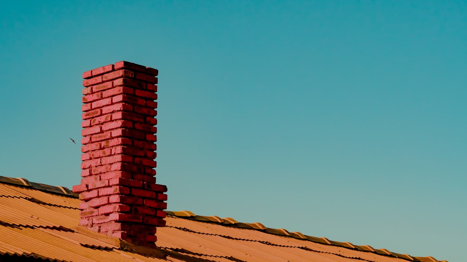 A red brick chimney rising from a terracotta tile roof against a teal sky, clean and free of people, text, or logos.