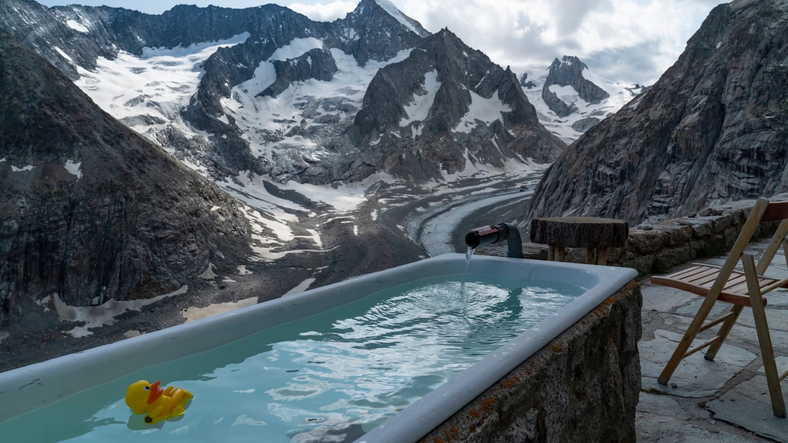 An outdoor cold-plunge tub filled with water and a yellow rubber duck, overlooking a dramatic snow-capped mountain range.