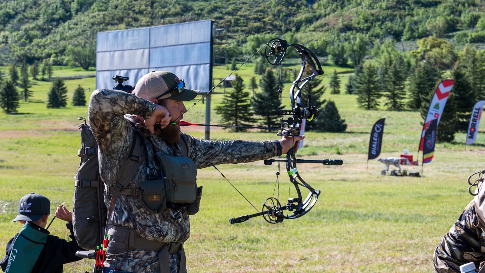 An archer drawing a compound bow outdoors in a grassy field.