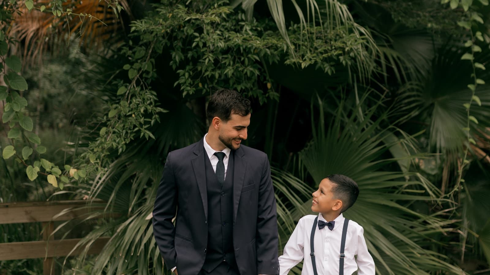 A father and young son in suits holding hands, walking outdoors in a lush garden.