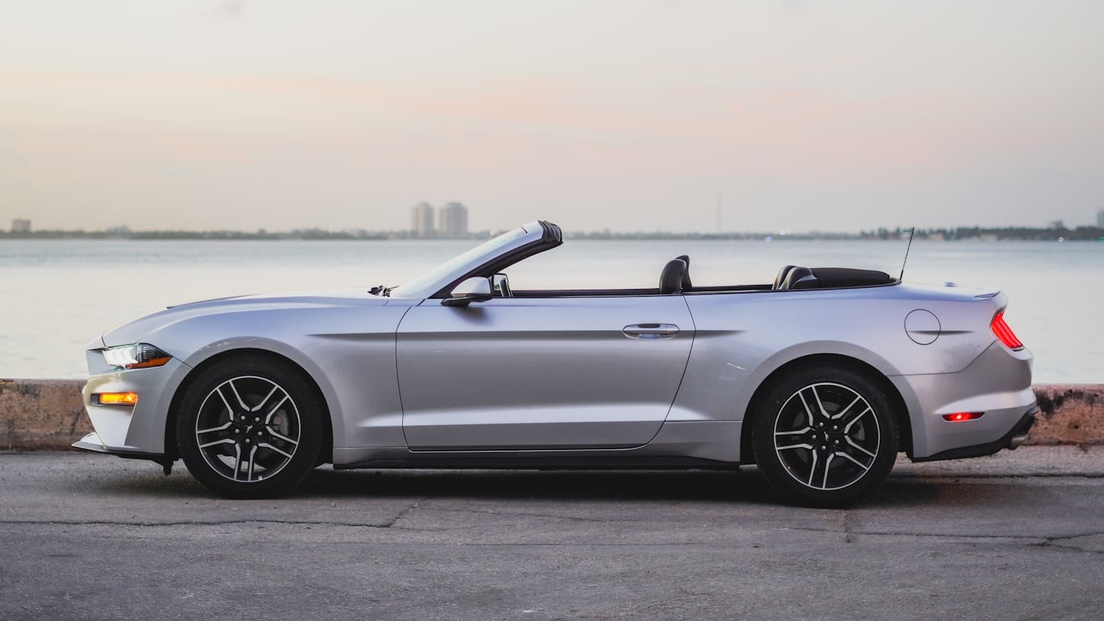 A silver convertible with its top down parked beside a waterfront at dusk, with no people, readable plate, or logo visible.