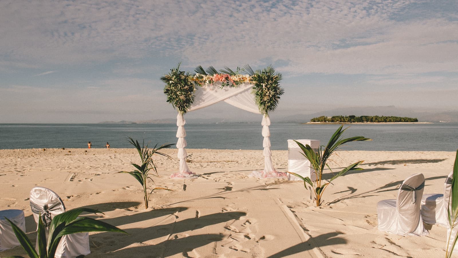 An empty white floral wedding arch set up on a tropical beach with the ocean and a small island in the background.