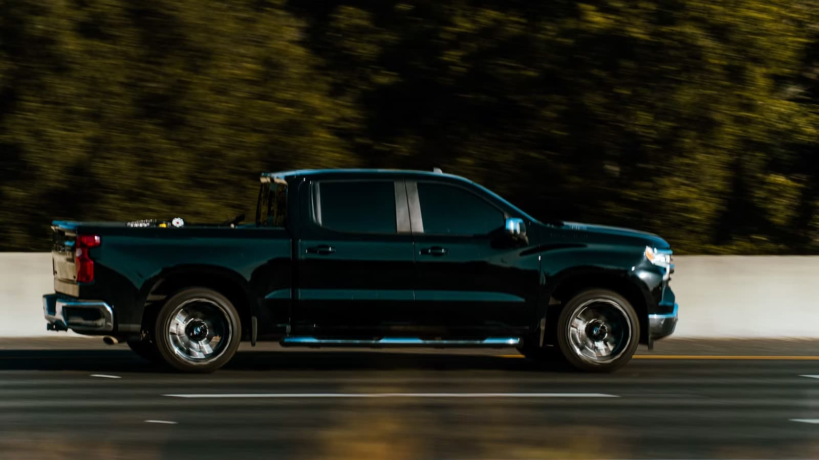 A dark modern crew-cab pickup truck in side-profile motion on a highway at night, with a blurred forested backdrop.