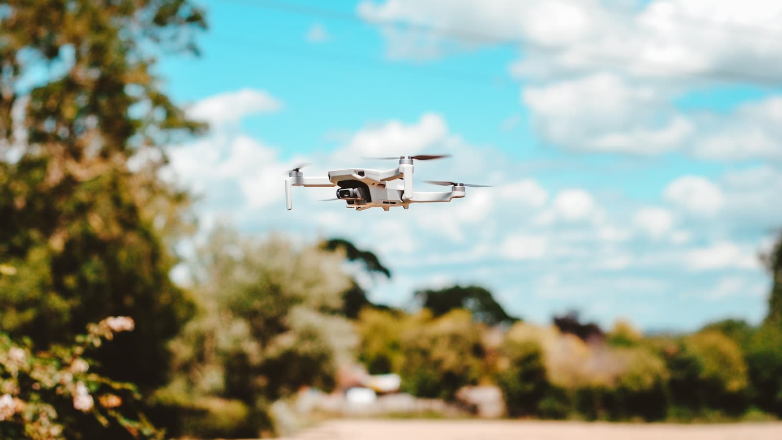 A small white consumer drone hovering against a blue sky with clouds and trees, with no readable branding visible.
