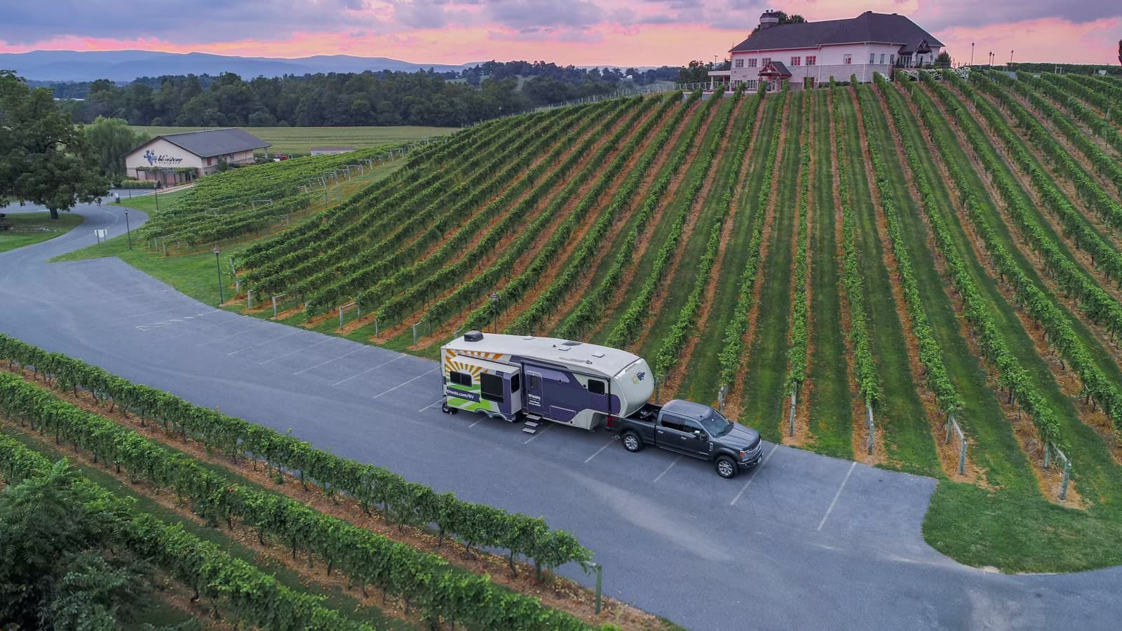Aerial view of a pickup truck towing a fifth-wheel trailer parked in a vineyard parking area at sunset.
