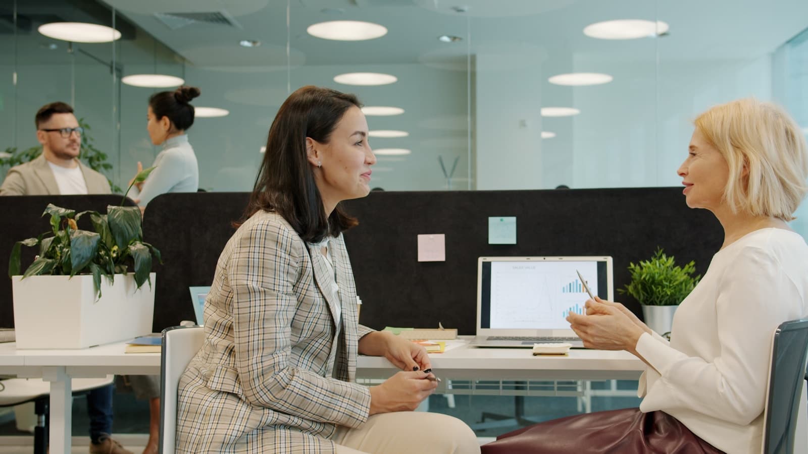 Two professional women in conversation at a desk, one gesturing at a laptop chart.