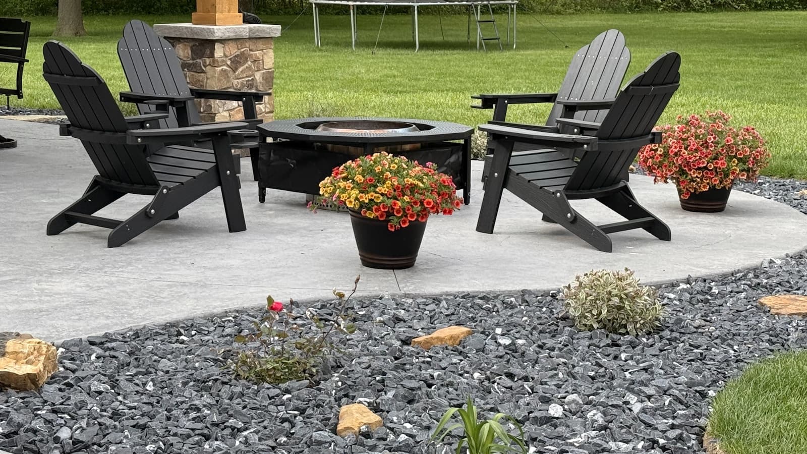 A backyard patio with black adirondack chairs around a metal fire pit, potted flowers, gravel landscaping, and a trampoline on the lawn behind.