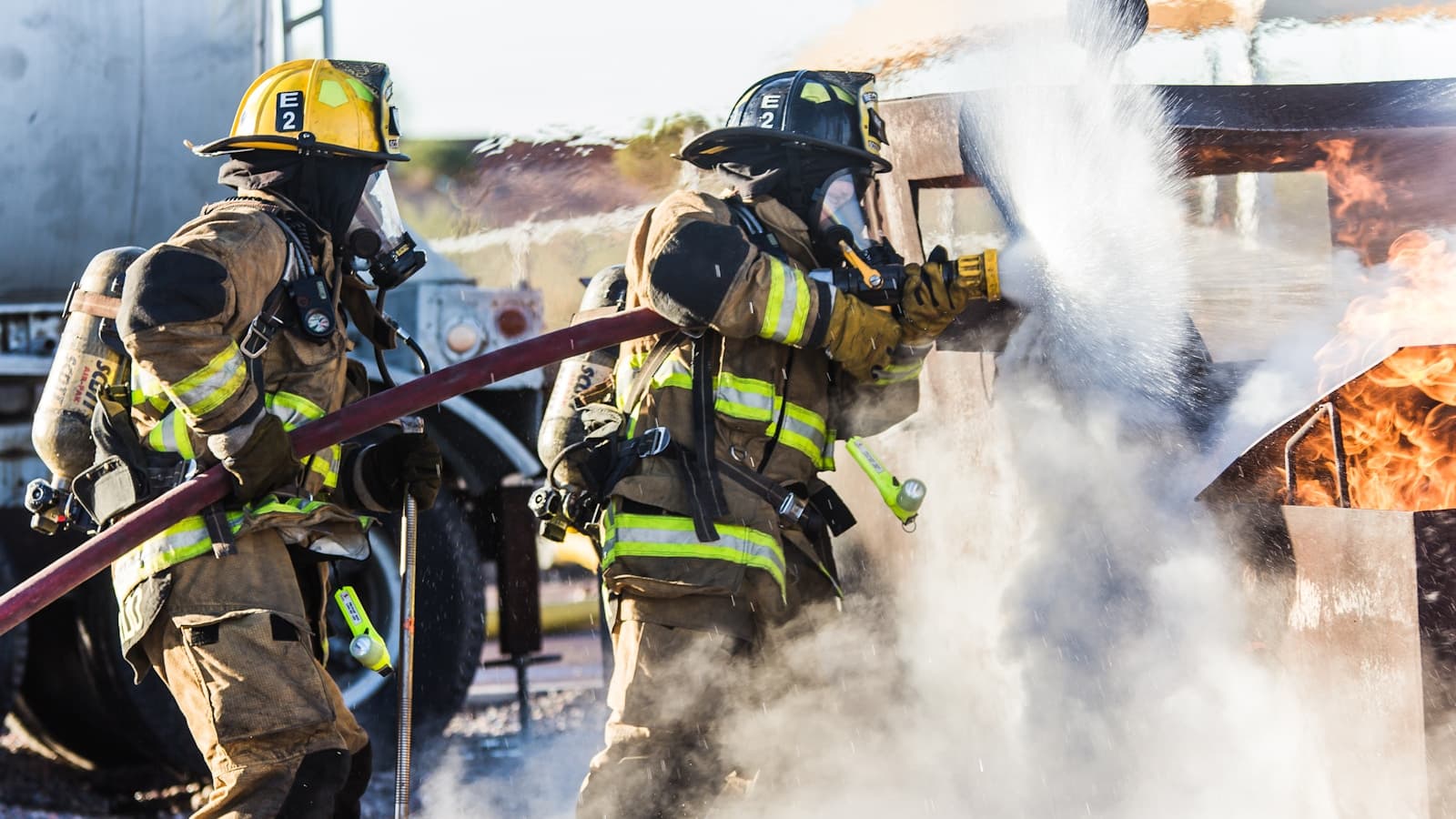 Two firefighters in turnout gear spraying water at a controlled training fire.