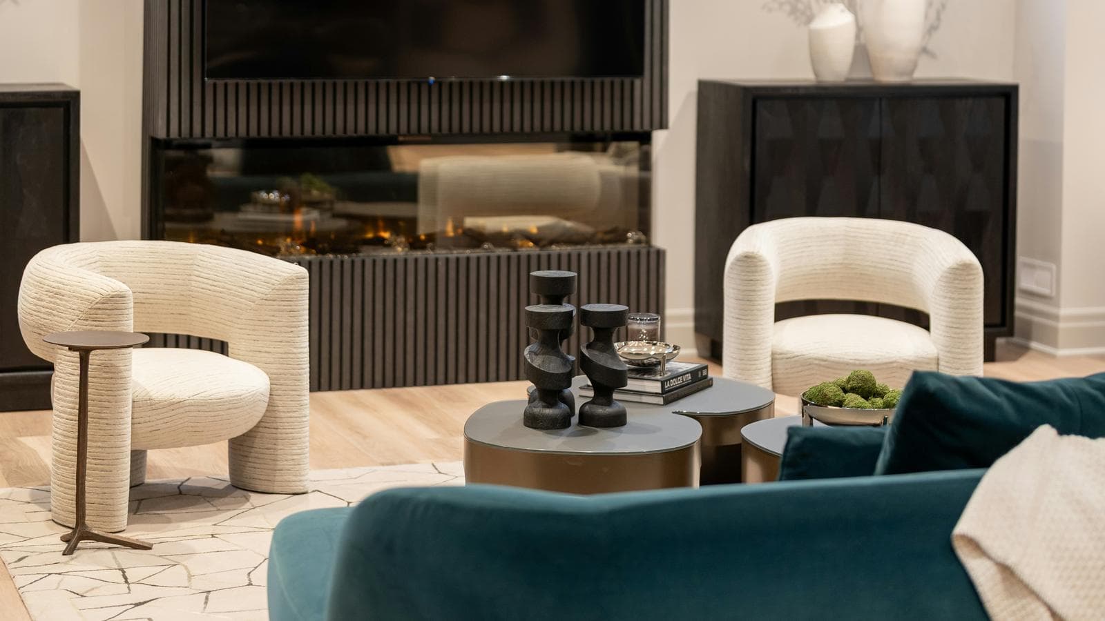 A modern living room with a wall-mounted TV above a built-in linear electric fireplace with visible flame, flanked by cream armchairs and a teal sofa.