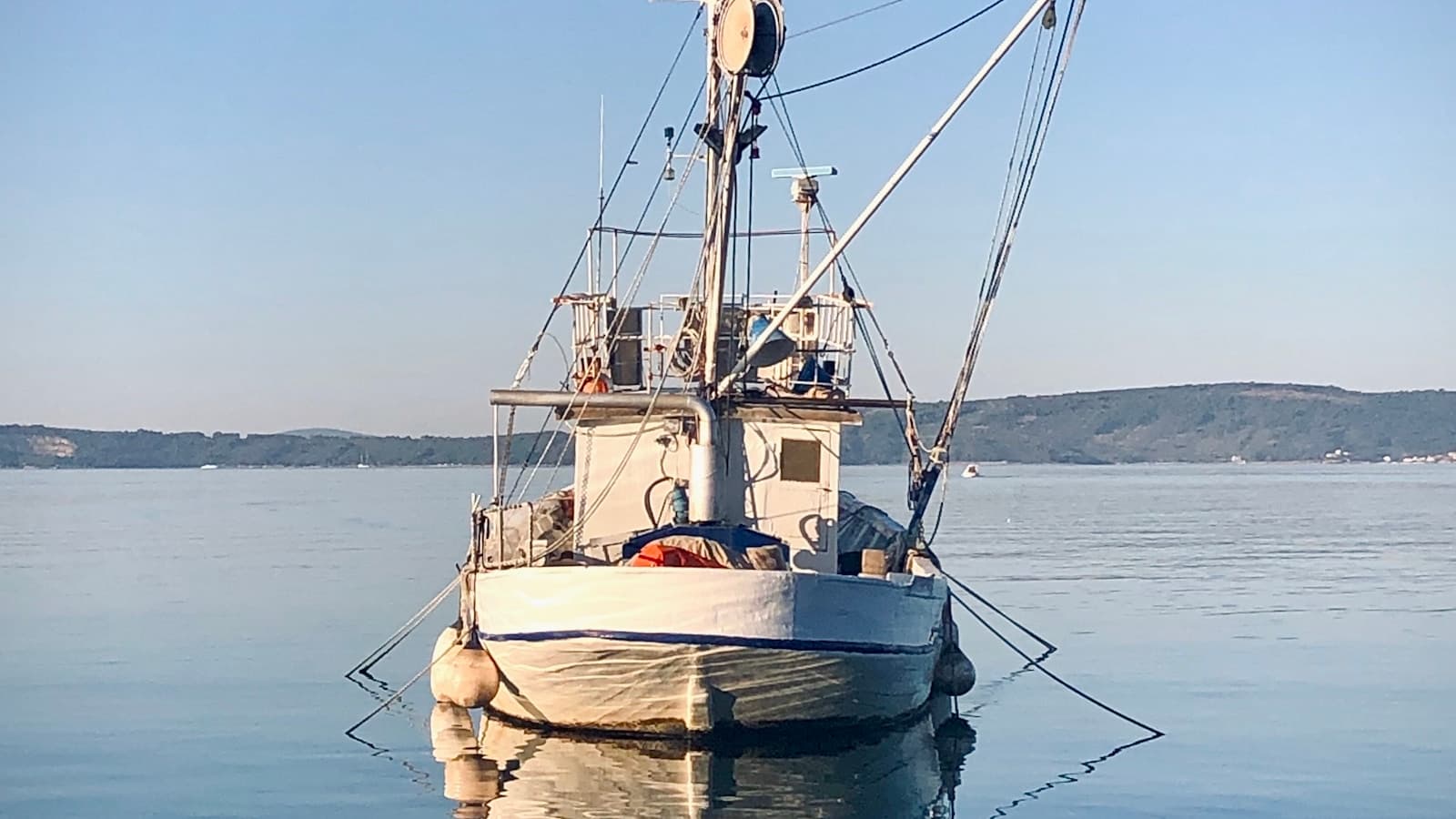 A small white fishing boat anchored on calm, glassy morning water.