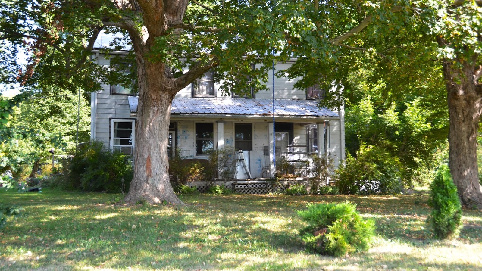 A weathered two-story farmhouse mid-renovation with peeling paint, exposed insulation board on the porch walls, and a worn front porch shaded by a large tree.