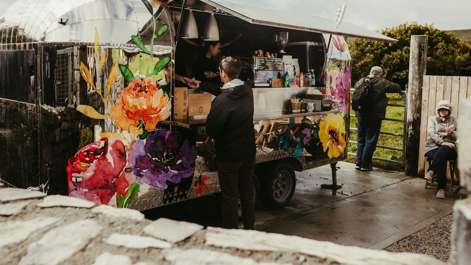 A silver food truck with a colorful painted flower design serving a customer at its open counter window.