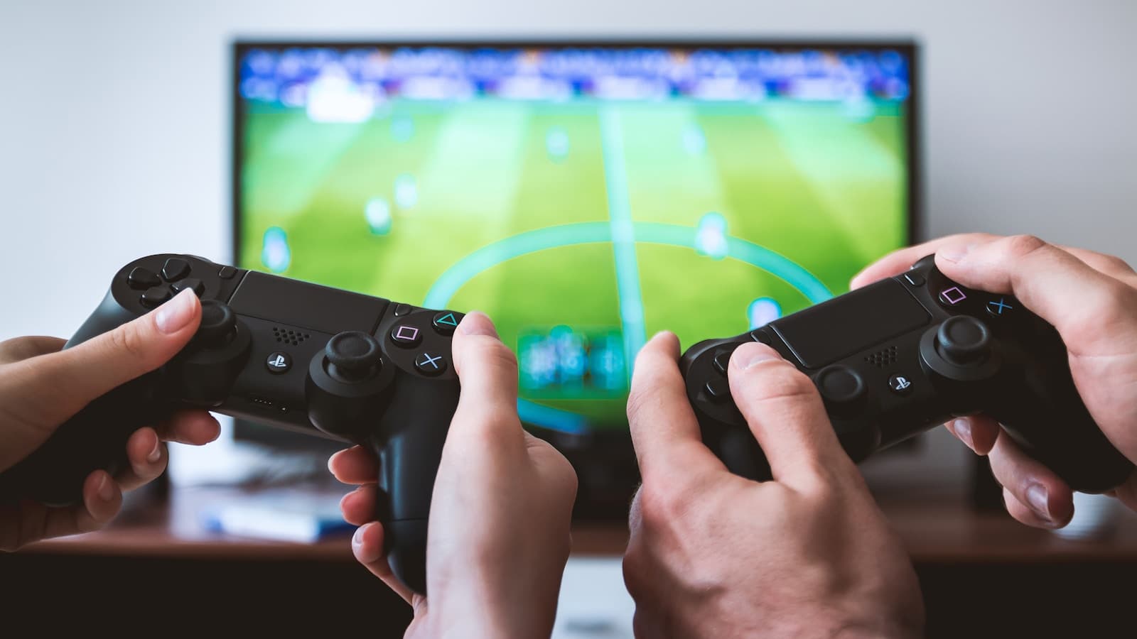 Two people's hands holding game controllers in front of a TV showing a blurred soccer video game, no readable text or logos.