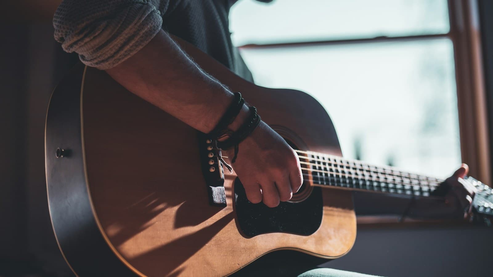 A person's hand strumming an acoustic guitar indoors near a sunlit window.